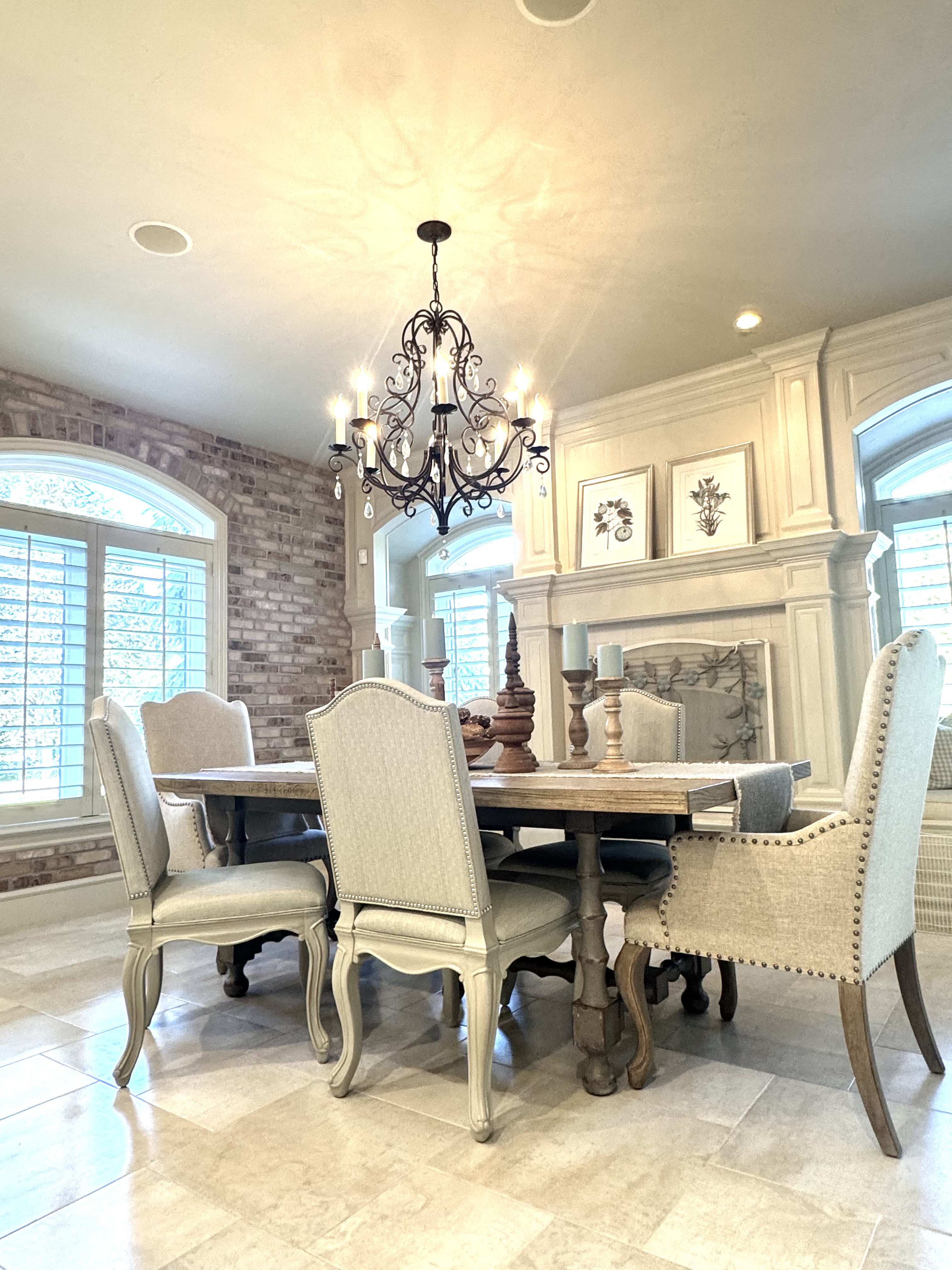 A dining room with an iron chandelier, upholstered chairs, exposed brick, and a paneled fireplace.